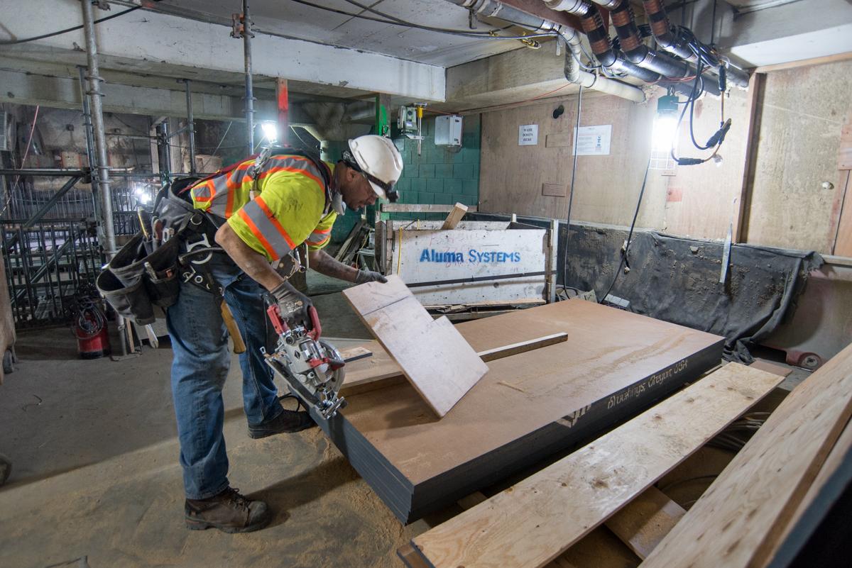 Worker in safety gear cutting plywood for concrete forms in transit station concourse.