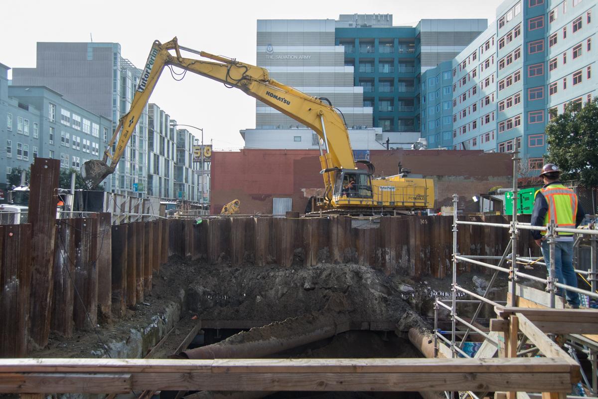 Excavator removing soil from underground station box at 4th Street construction site with workers and steel sheet piling visible.
