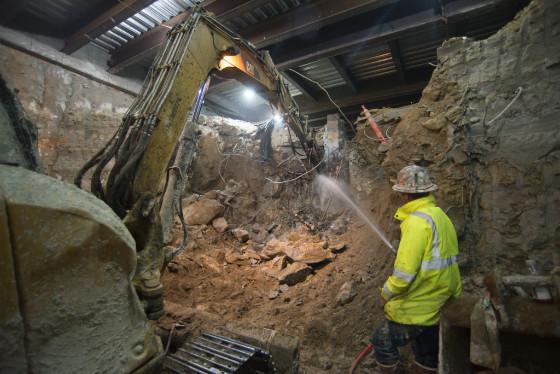 Excavator with jackhammer breaks up soil while worker in yellow vest sprays water to control dust at construction site.