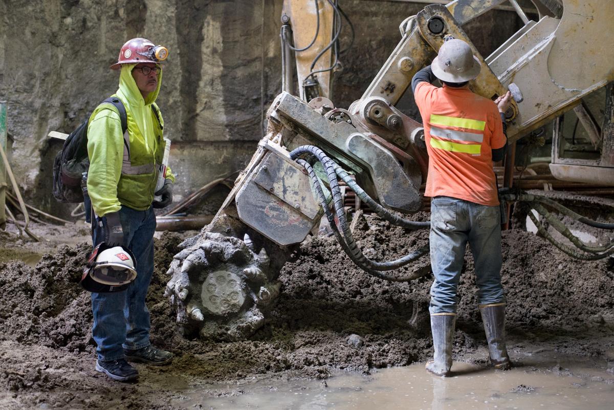 Two construction workers in safety gear operate a concrete pump truck during underground station excavation work.