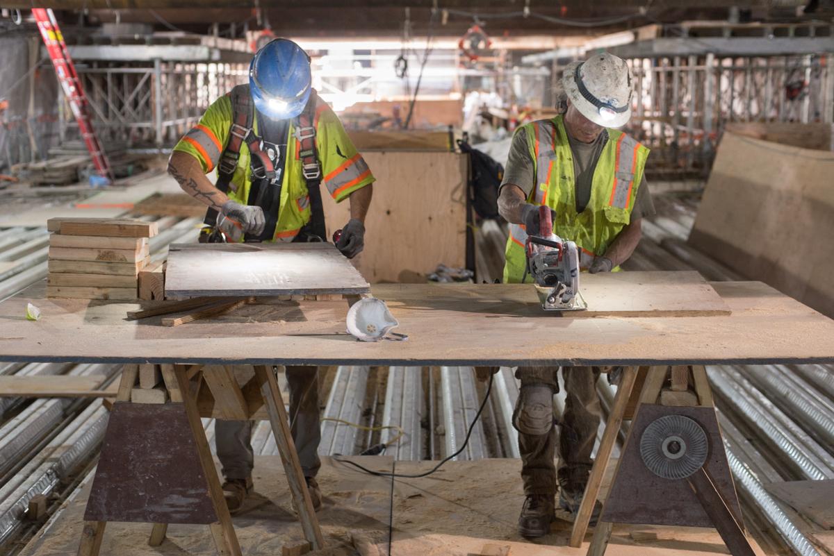 Two construction workers in safety gear cut plywood at a worksite on a mezzanine level.