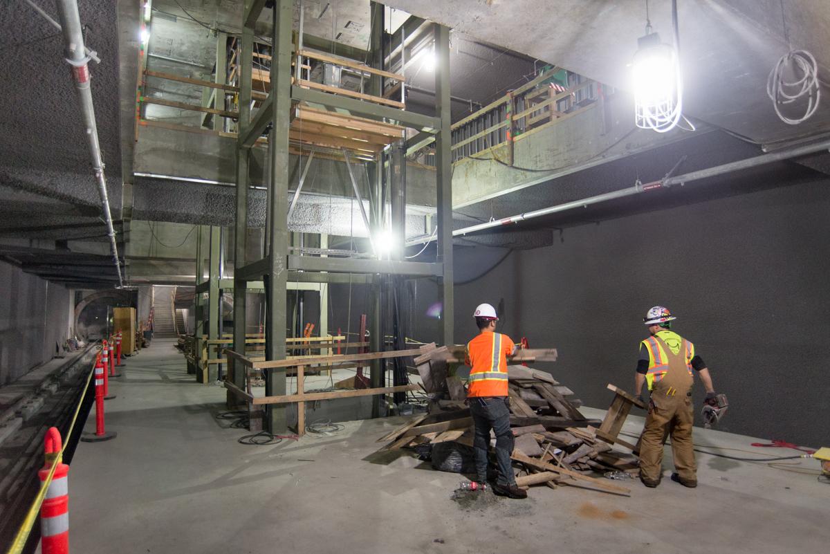 Workers in safety gear inside underground transit station under construction with structural framework and equipment visible.