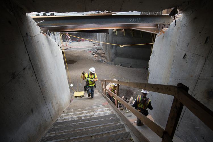 Workers on temporary bridge inside underground transit station construction site with concrete walls and steel beam overhead.