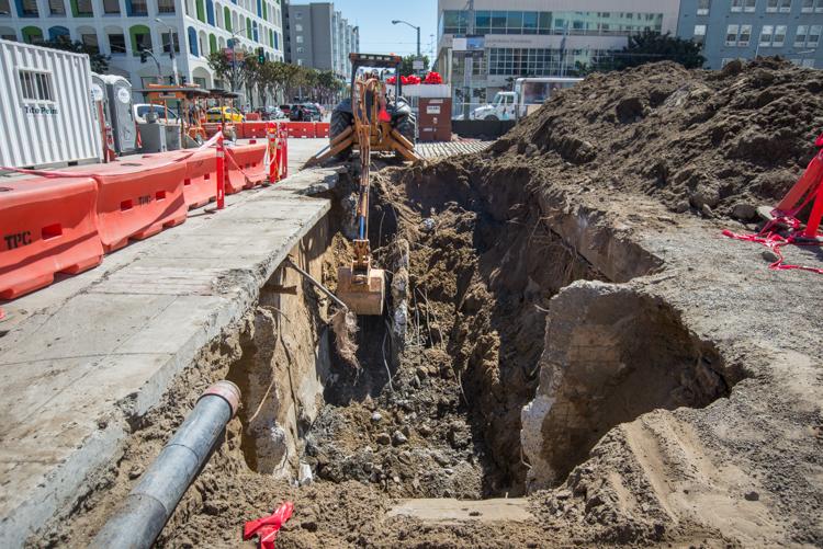 Backhoe excavating concrete and soil in urban construction site with red barriers and buildings in background