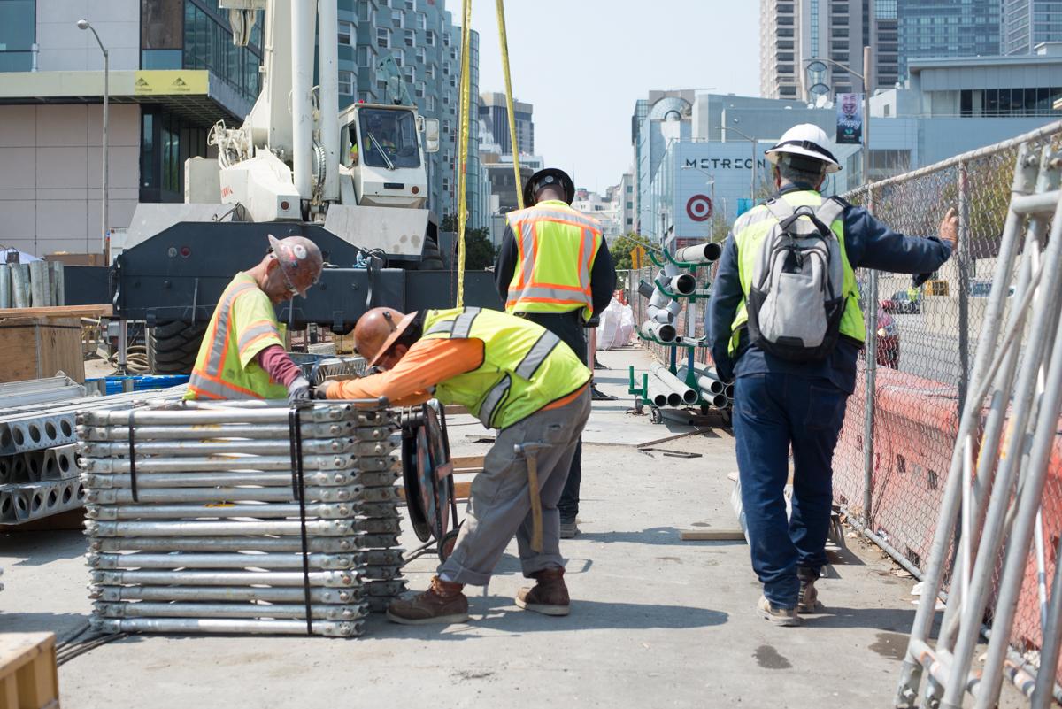 Construction workers in safety vests organize metal barriers and equipment on an urban street staging area.