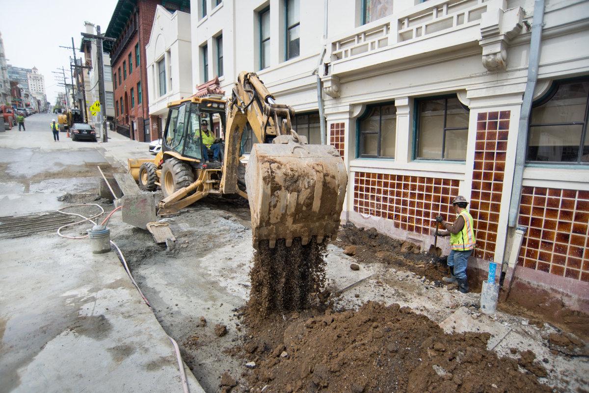 Excavator removing soil from street excavation site with construction worker nearby and historic buildings in background.