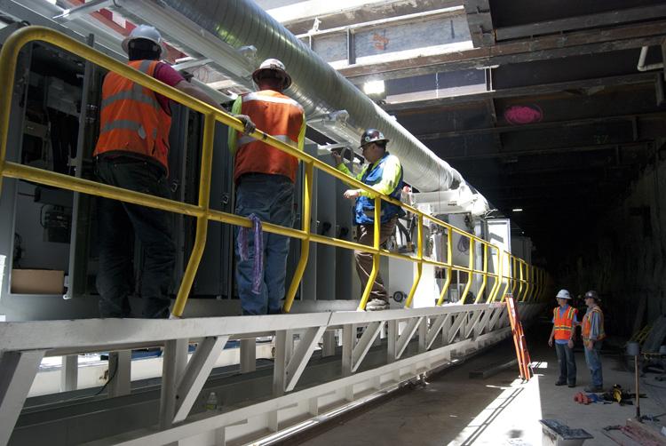Construction workers in safety vests assemble tunnel boring machine components on a platform in an underground tunnel.