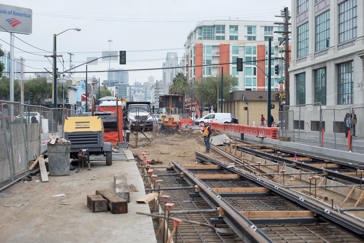 Construction site with track installation work, heavy equipment, and red safety barriers along urban street.