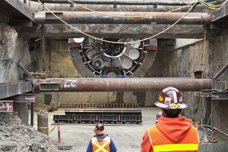 Tunnel boring machine ring being lowered into underground transit tunnel with workers observing below.