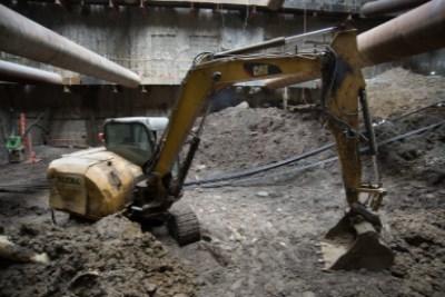 Excavator operating at underground transit construction site with concrete structure overhead and construction materials visible.