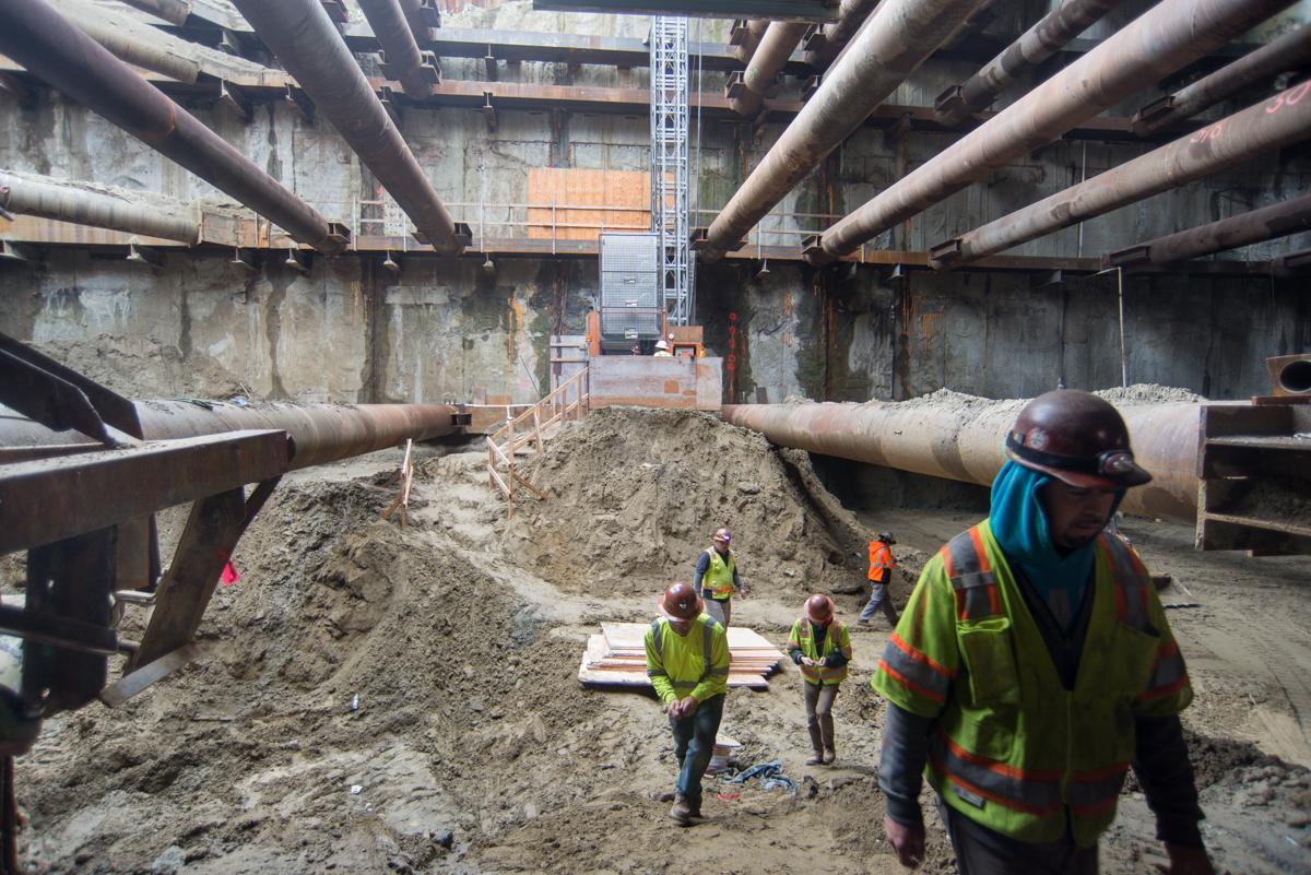 Construction workers in safety vests navigate excavation site beneath large industrial pipes at transit station construction.