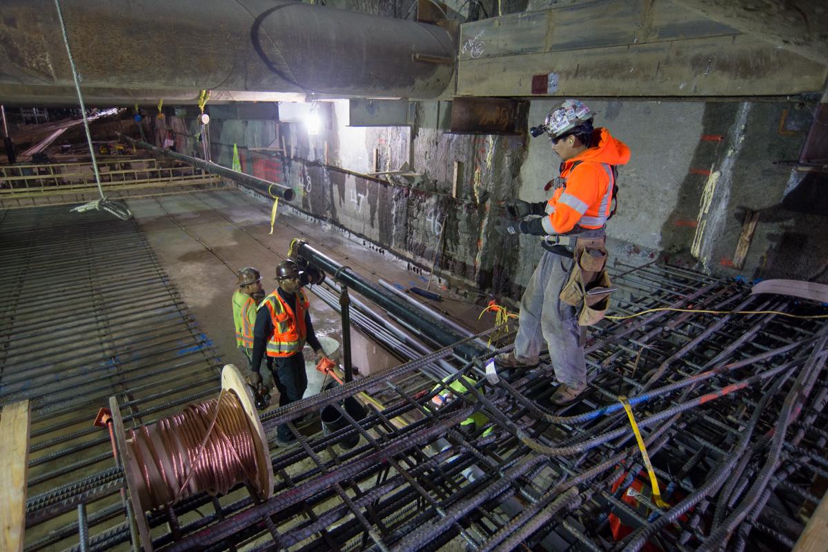 Workers in safety vests assemble rebar reinforcement cage for station box construction underground.