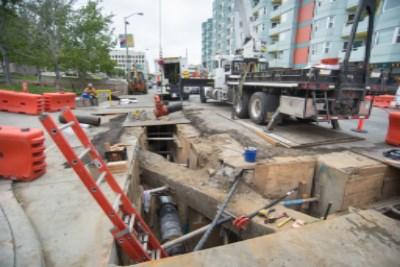 Construction site on 4th Street with excavated trench, concrete work, and delivery truck among red barriers.