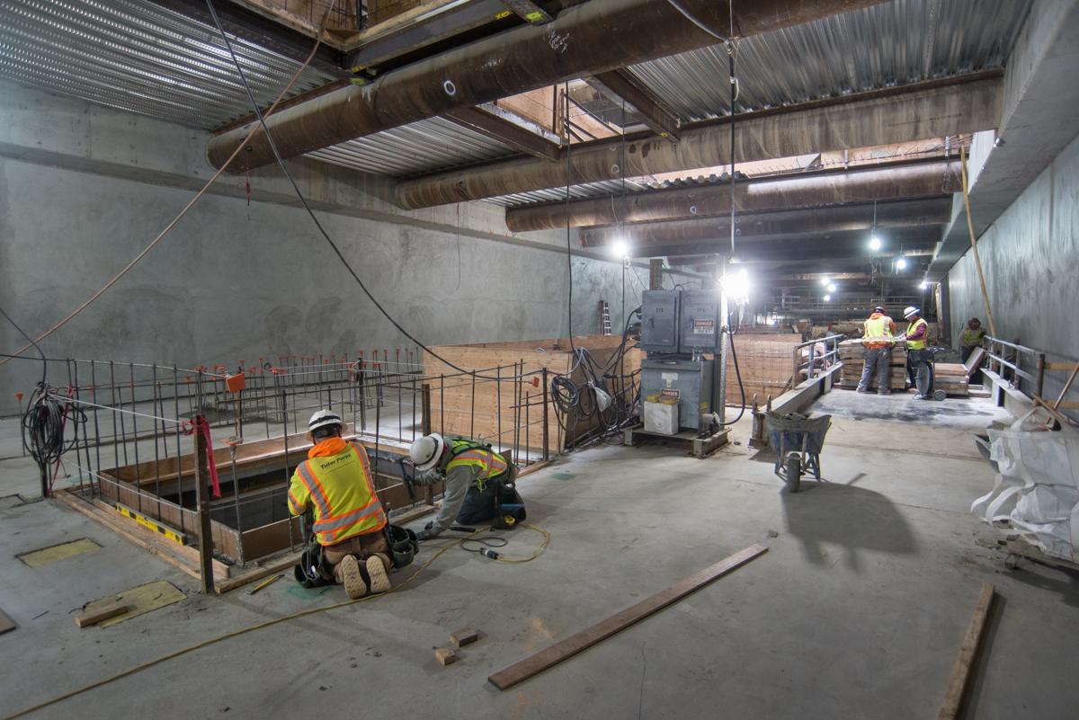 Construction workers in safety vests work on concrete formwork inside a transit station mezzanine level under industrial lighting.