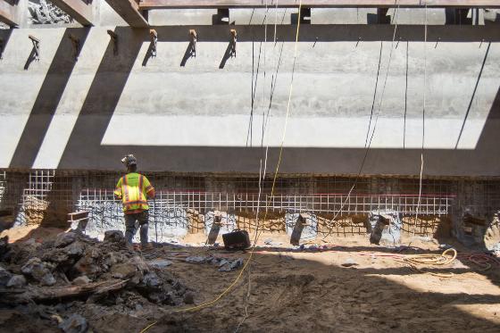 Construction workers installing reinforcement mesh on station wall beneath concrete structure