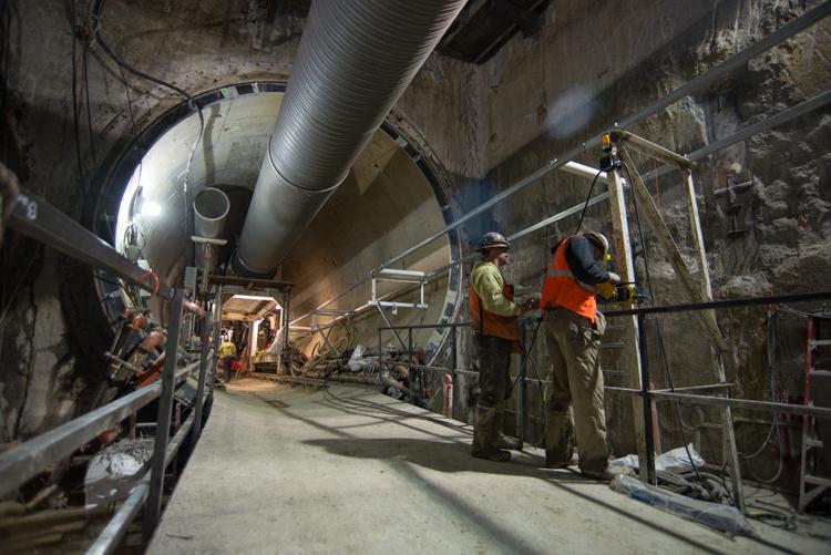 Workers in safety vests operate conveyor system inside underground tunnel construction site.