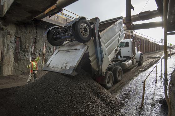 A worker in a safety vest directs a dump truck unloading gravel material at a construction site beneath a concrete structure.