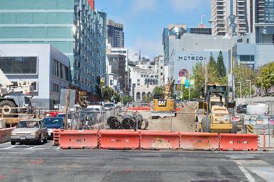 Urban street construction site with excavators, orange barriers, and city buildings in background.