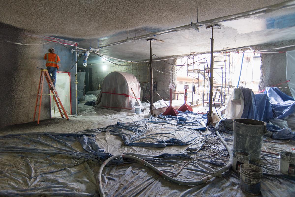 Interior construction work at transit station with protective sheeting, scaffolding, and worker on ladder.