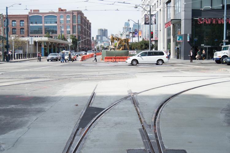 Streetcar tracks under construction at intersection with urban street, buildings, and vehicles visible.