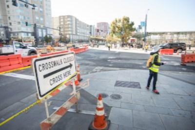 Worker in yellow vest marking street intersection with crosswalk and sewer work barriers visible.