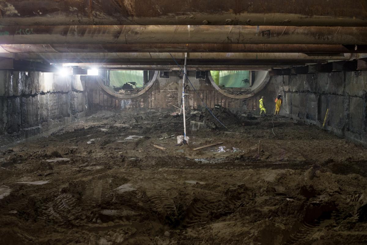 Underground construction site with excavated earth, concrete walls, support columns, and workers in safety vests under industrial lighting.