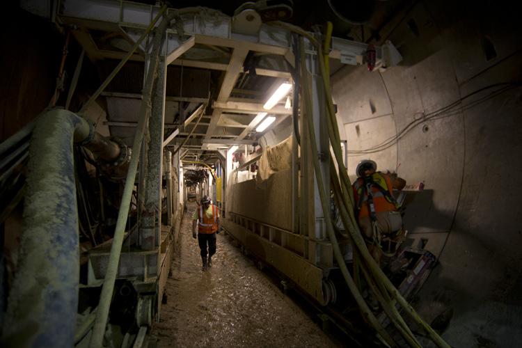 Workers walk through underground tunnel construction site with exposed pipes and support structures overhead.