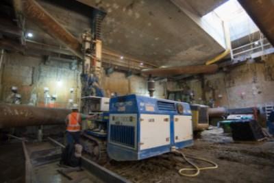 Industrial drill rig and equipment inside deteriorating concrete station headhouse with temporary steel supports.