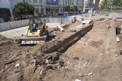 Excavator operating in urban construction site with sheet pile trench and demolished garage debris visible.