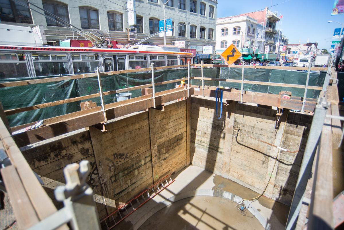 Construction site showing excavated shaft with concrete walls, steel beams, and safety fencing on Stockton Street.