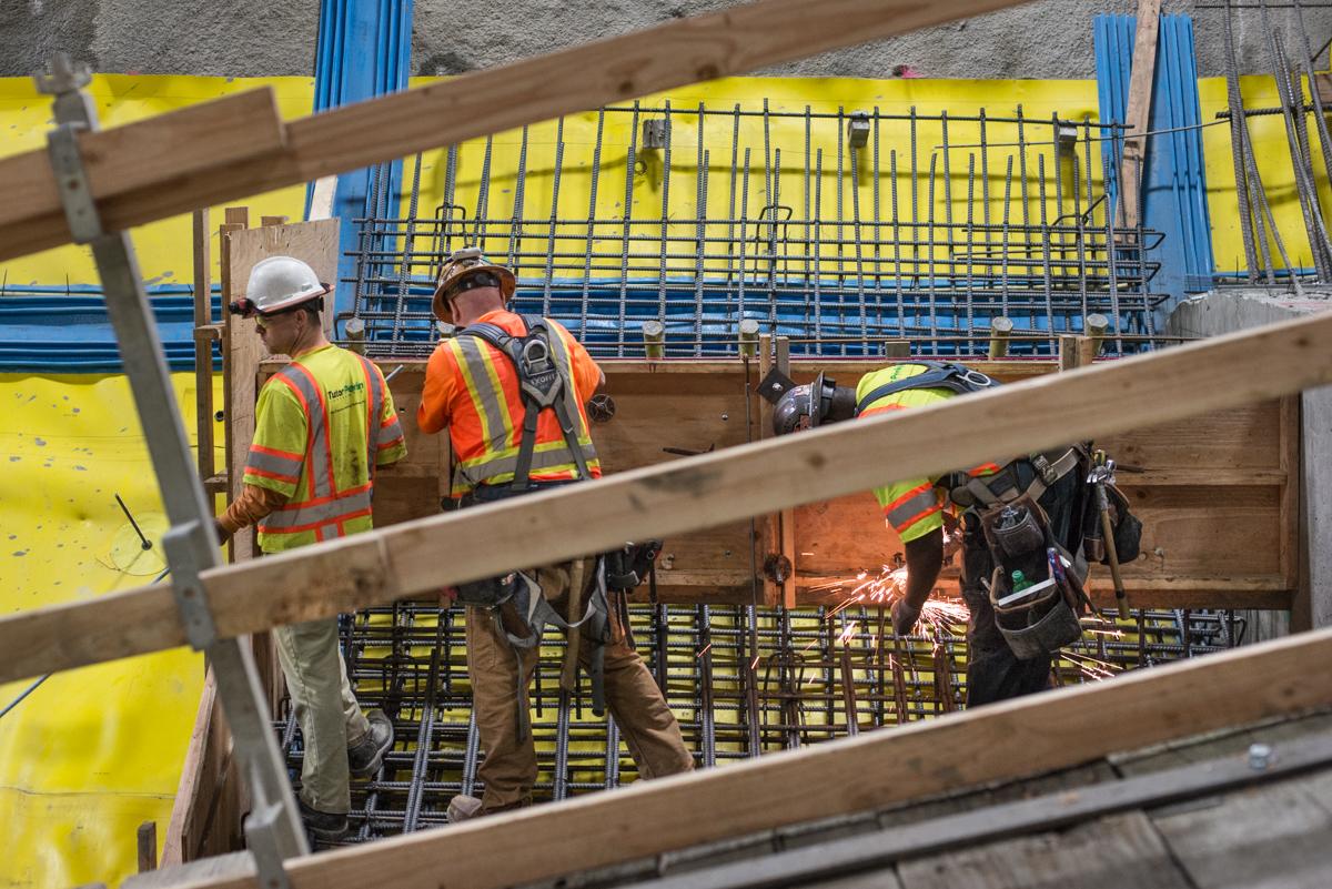 Construction workers in safety gear install rebar in underground station cavern with yellow shoring and blue support beams.