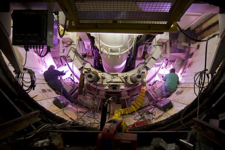 Workers weld tunnel boring machine Mom Chung's shield in underground construction site with purple lighting.