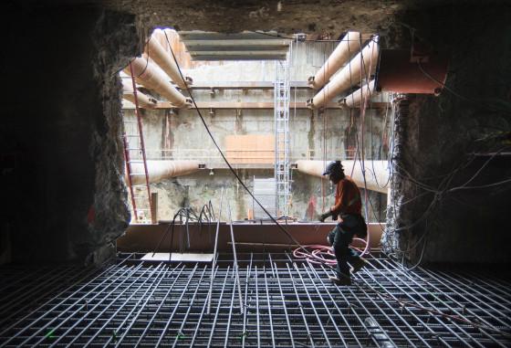 Worker on rebar framework inside underground transit station construction site with headhouse opening visible above.