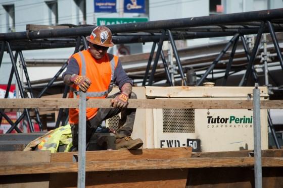 Construction worker in safety gear operating equipment on wooden platform above Stockton Street excavation site.