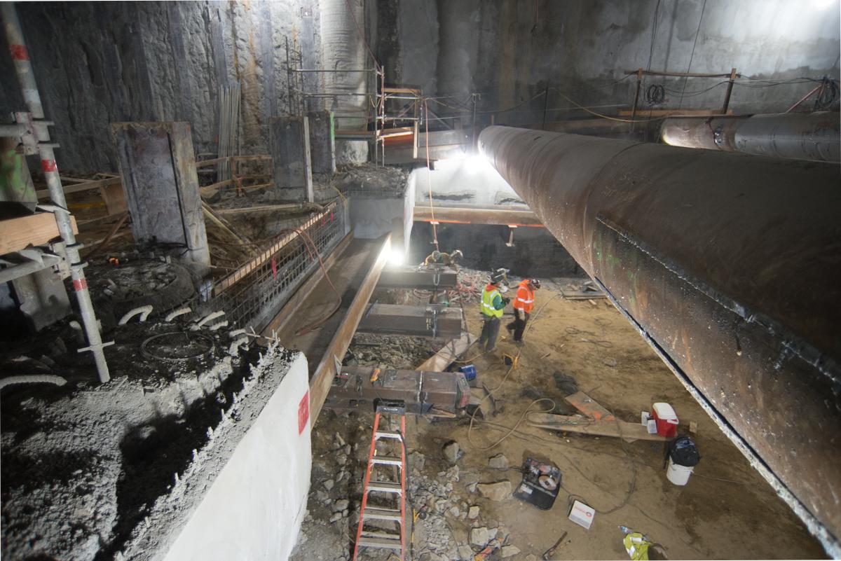 Workers in safety vests install temporary bracing inside an underground transit station construction site.