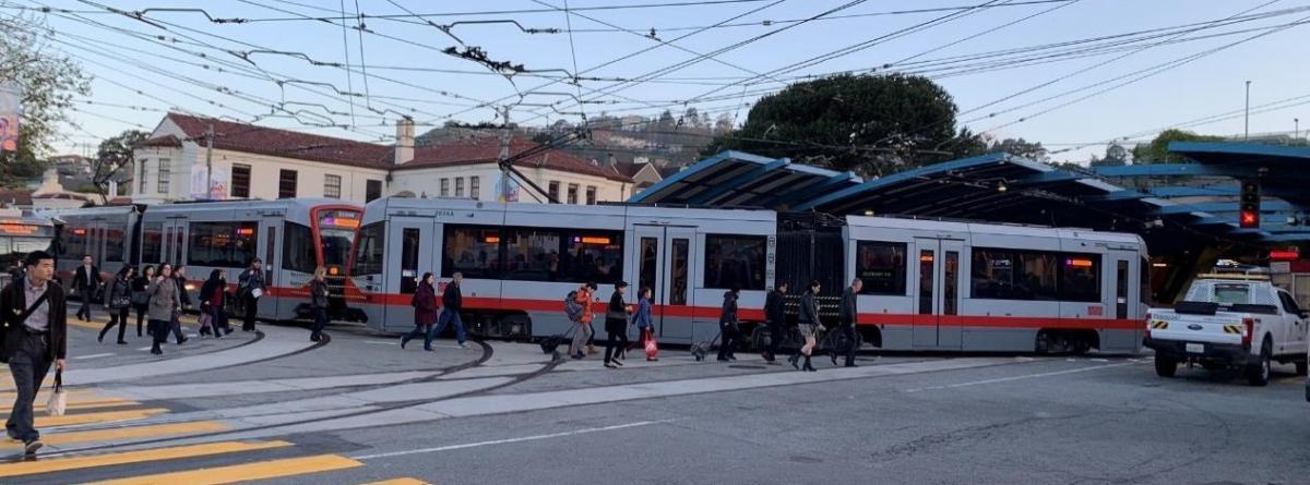 Red and white light rail train at West Portal Station with pedestrians crossing nearby.