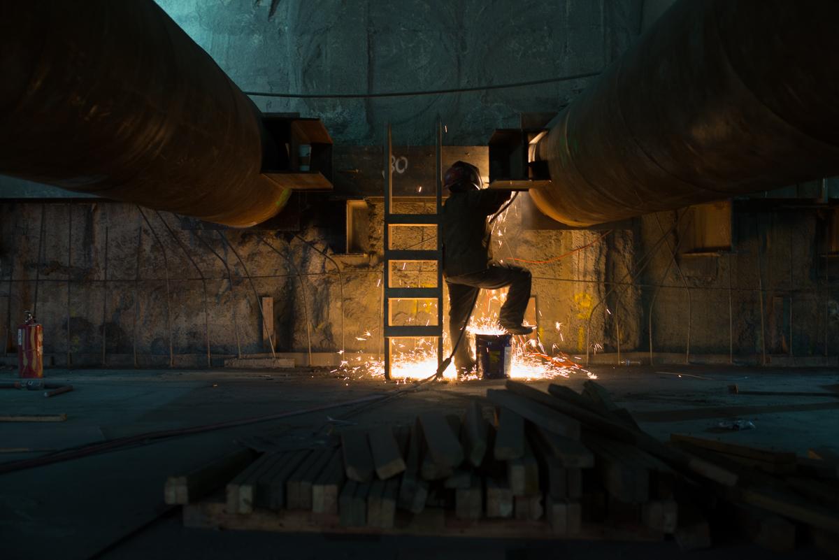 Worker welding metal brackets inside underground transit station construction area with temporary support beams.