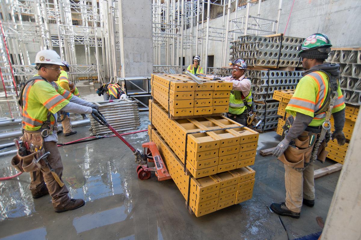 Construction workers unpack and organize yellow metal scaffolding components inside a transit station headhouse.
