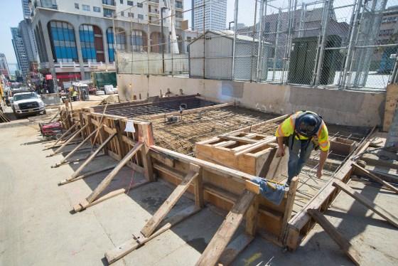 Worker in yellow safety gear installing wooden forms and rebar for concrete pads at Central Subway construction site.
