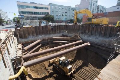 Construction excavation site with steel sheet piling, large pipes, and yellow bulldozer removing material from underground station.