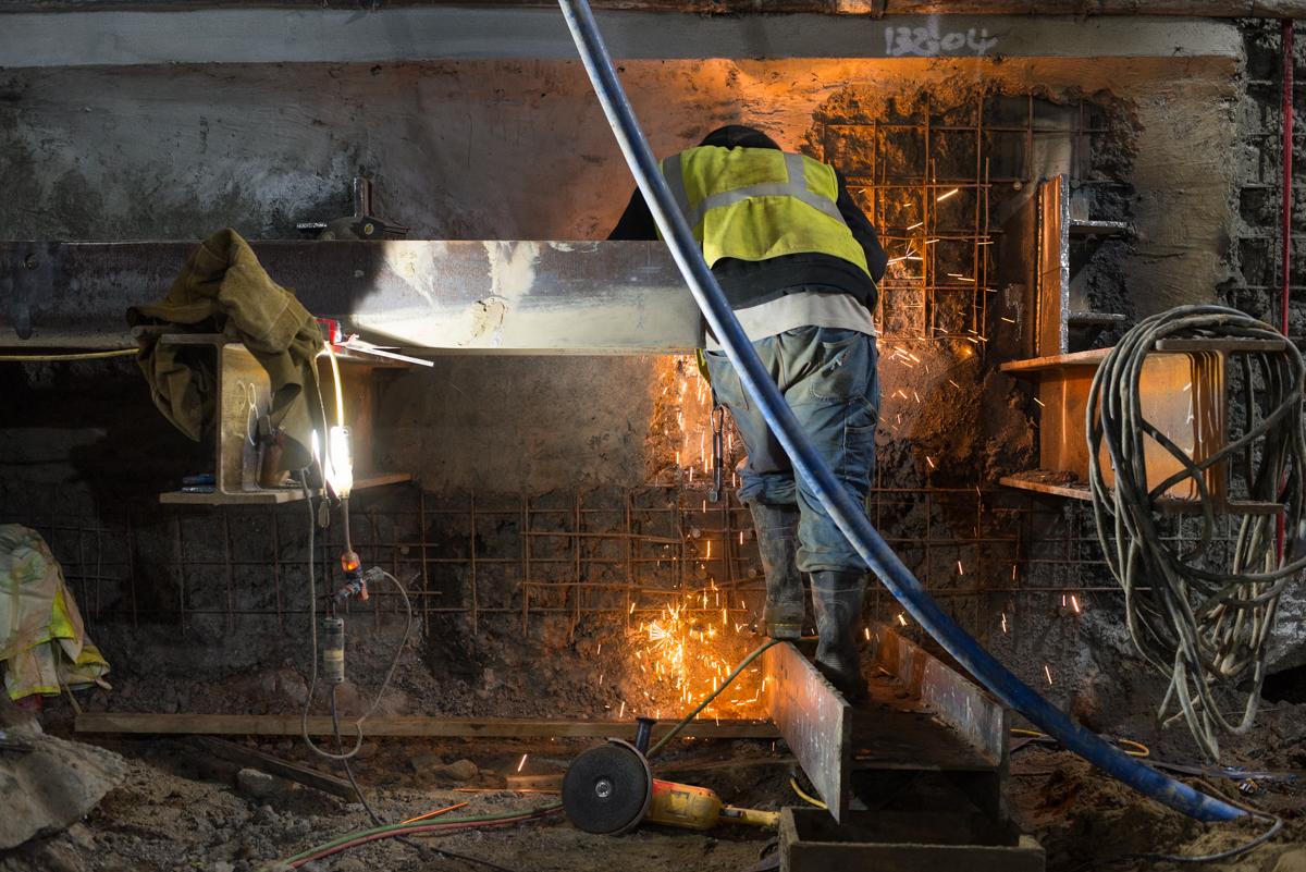 Worker in safety vest welding steel beam inside underground station construction area with sparks flying.