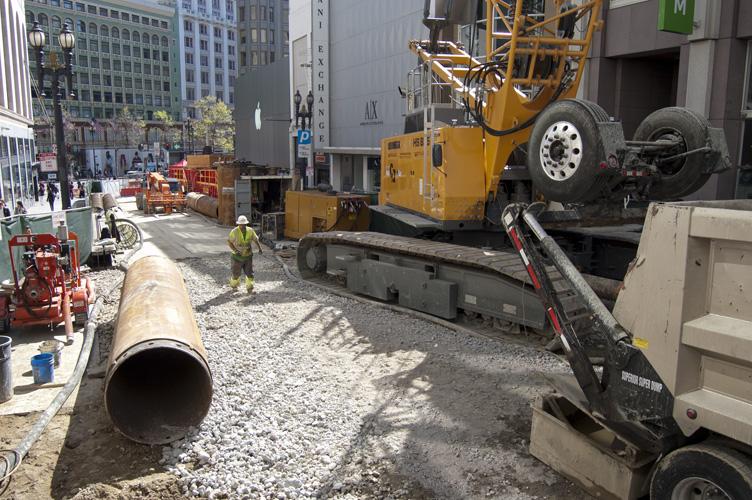 Construction crew operating concrete pump truck on urban street with large pipe and equipment.