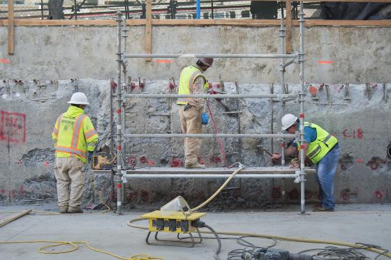 Construction workers in safety vests remove concrete from a wall section using tools and equipment, exposing rebar underneath.