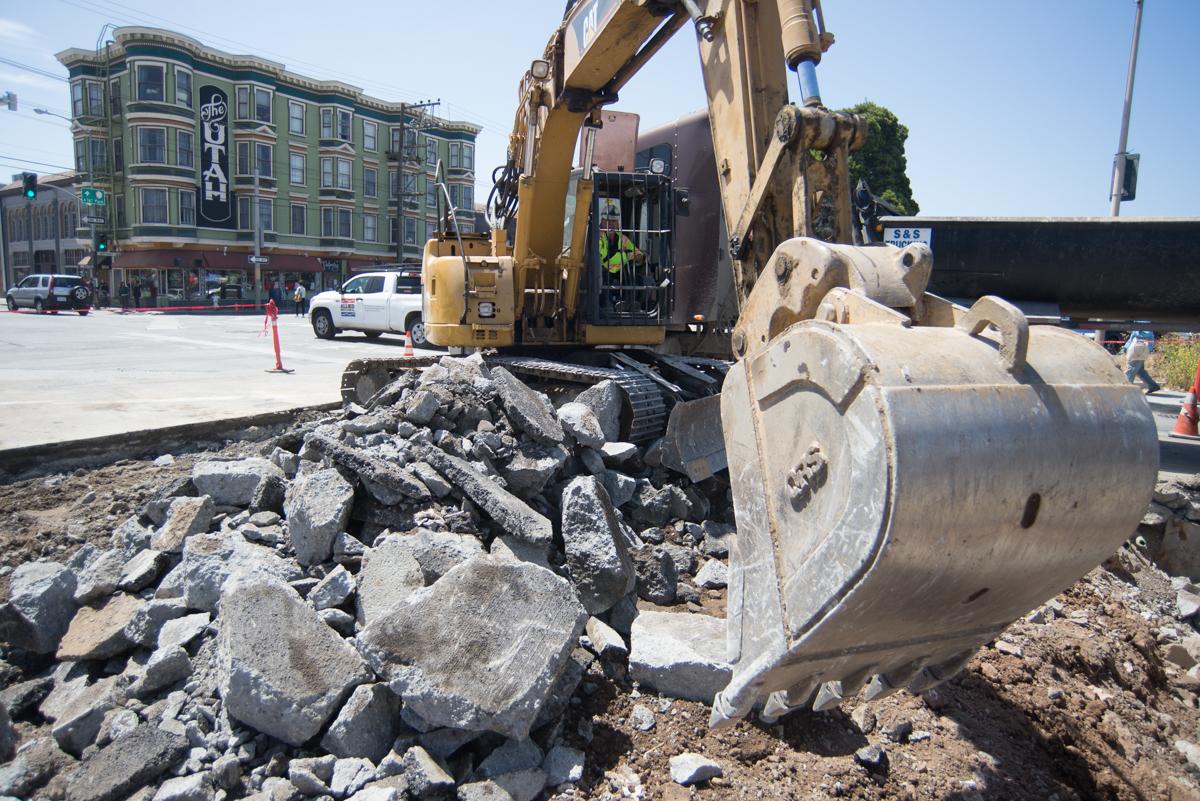 Excavator with bucket sorting concrete debris on city street during Central Subway construction.