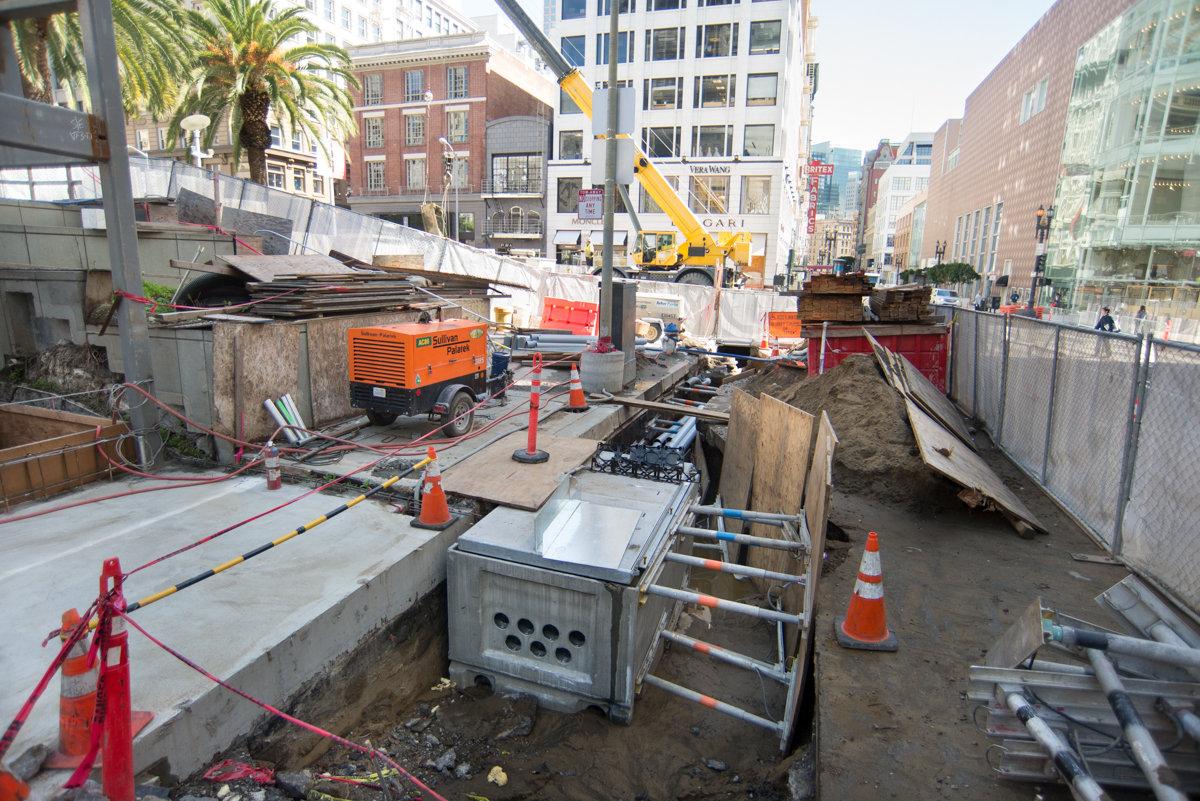Construction site on Ellis Street with excavation pit, orange compressor, crane, and surrounding urban buildings.