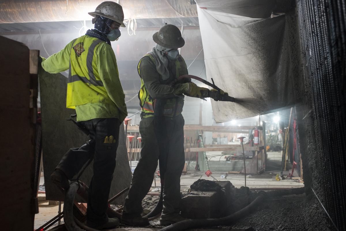 Two workers in hard hats and safety vests spray shotcrete on an underground concrete wall during transit construction.