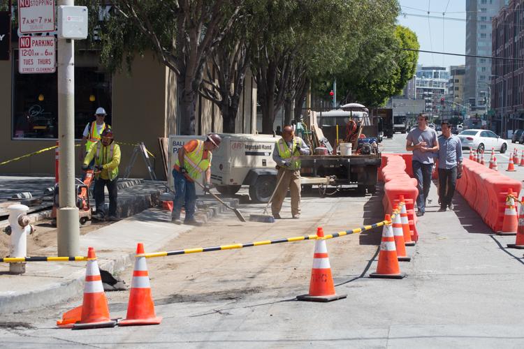 Construction workers in safety vests clean up a street work zone surrounded by orange traffic cones and barriers.