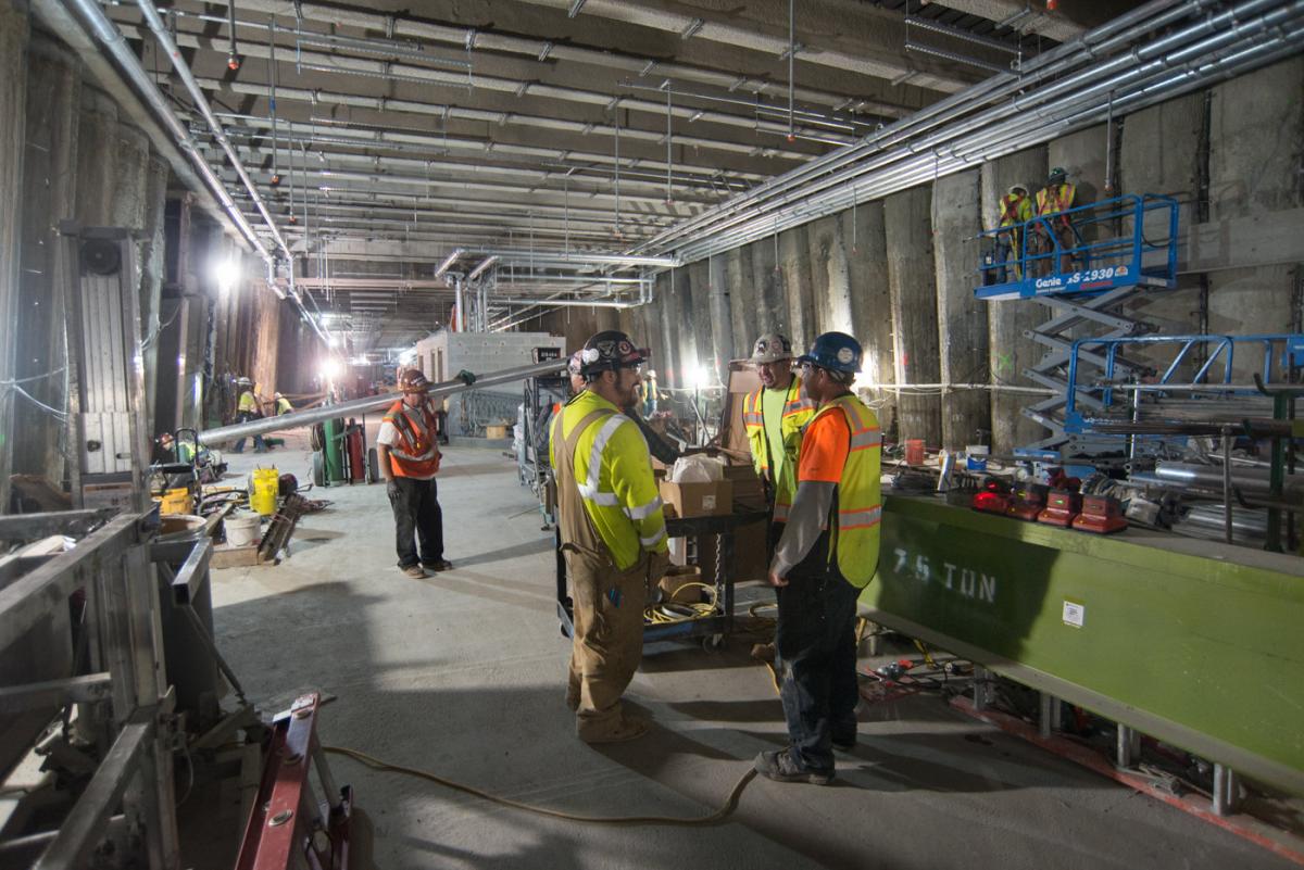 Construction workers in safety vests gather on mezzanine level installing utility conduits at Union Square/Market Street Station.