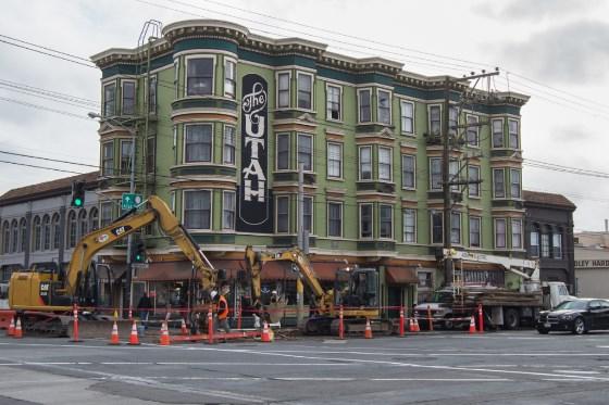 Historic green building with vertical "UTAH" sign undergoing construction with yellow excavators and equipment on street.