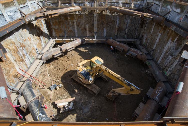 Aerial view of construction shaft with yellow excavator removing soil from wooden-lined underground pit.